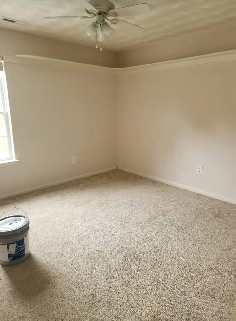 Bedroom in the original cream with white crown molding and a paint bucket on the carpet