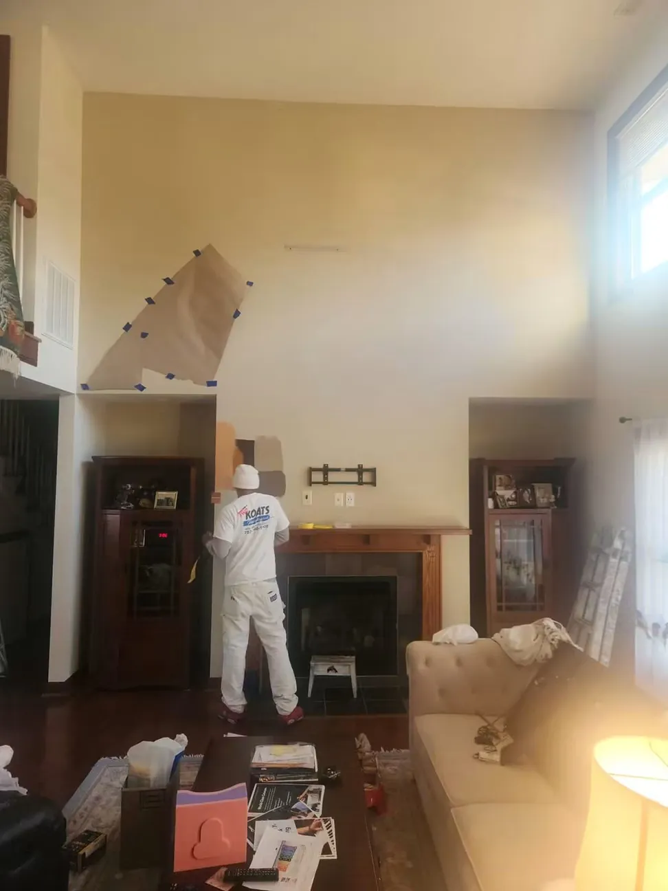 A painter in whites cutting in the chimney breast of an occupied great room, the furniture pulled back and sheeted