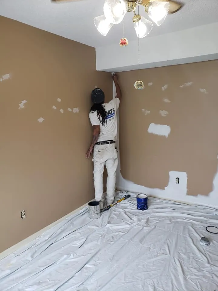Painter in branded shirt cutting in white ceiling line above patched tan bedroom walls