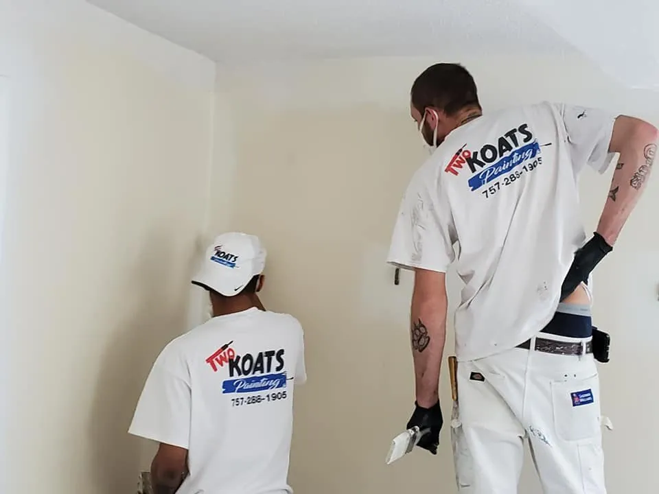 Two painters in Two Koats Painting t-shirts prepping a cream interior wall, one wearing a dust mask