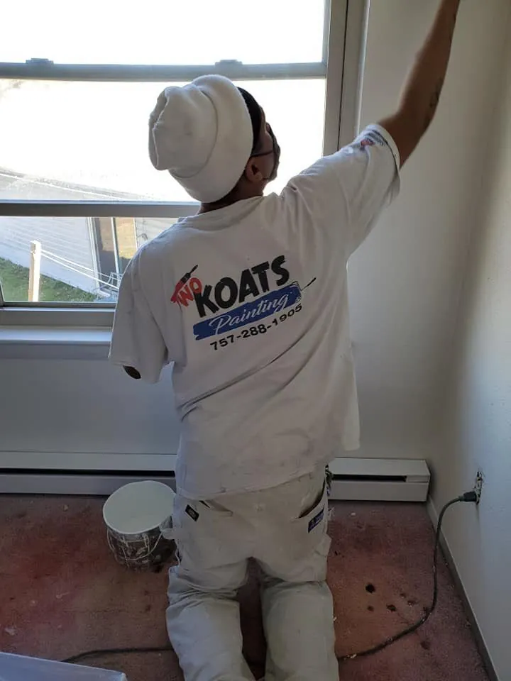 Painter in a Two Koats Painting t-shirt cutting in white paint along a wall beside a window