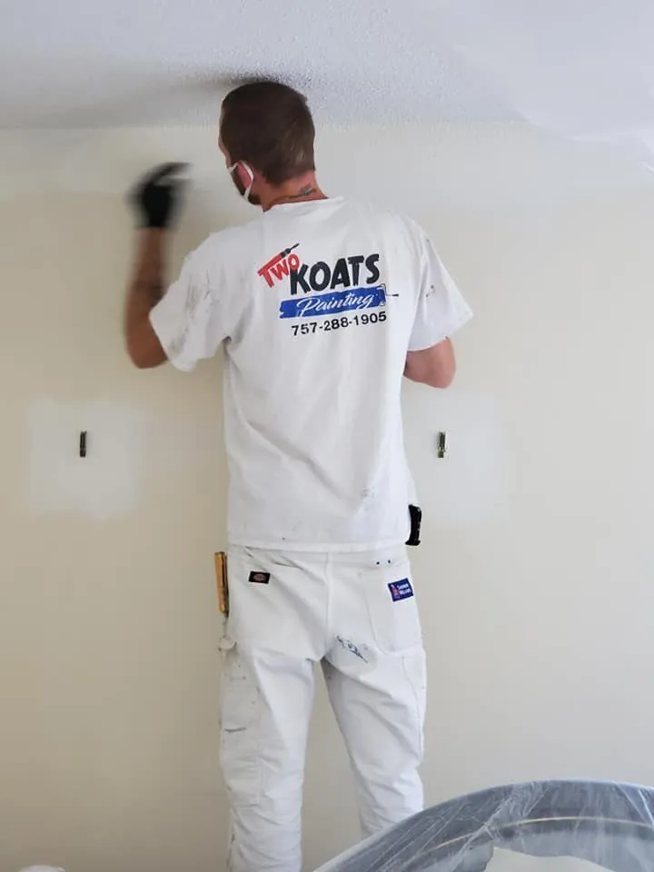 Painter in a Two Koats Painting shirt and white work pants working along a wall and ceiling line