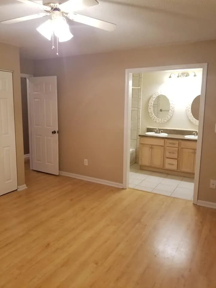Empty bedroom with tan walls and laminate flooring opening to a bathroom with double vanity