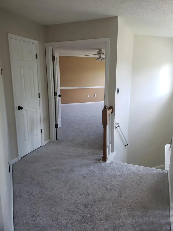 Upstairs hallway with cream walls, white six-panel door and gray carpet looking into a peach-walled room