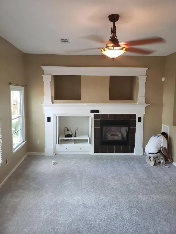 Living room with white fireplace mantel and built-in surround, tan walls, and worker painting a baseboard