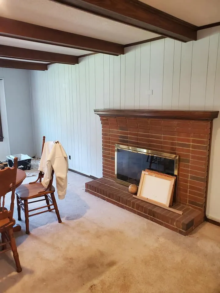 Vertical wood paneling painted white beside brick fireplace with dark stained ceiling beams